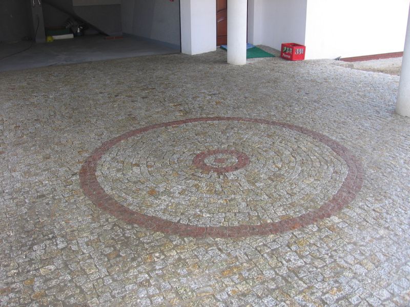 Podjazd wyłożony drobną kostką granitową w odcieniach szarości, z dekoracyjnym wzorem koła w kolorze czerwono-brązowym, otoczony kolumnami i zadaszeniem.